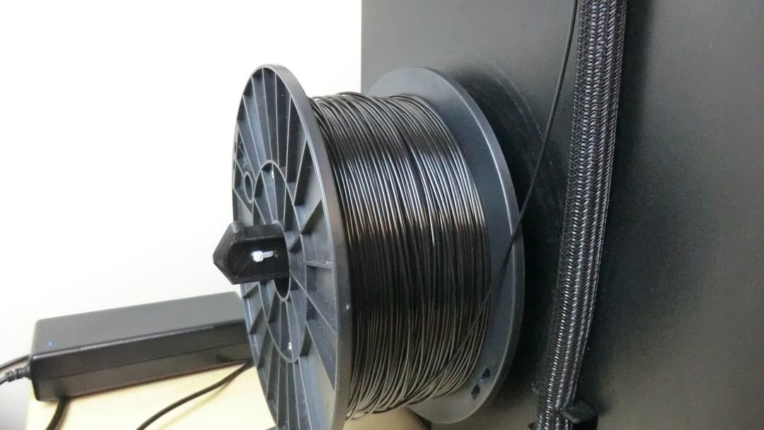 A spool wound with black 3D printer filament mounted on the side of a dark printer frame.