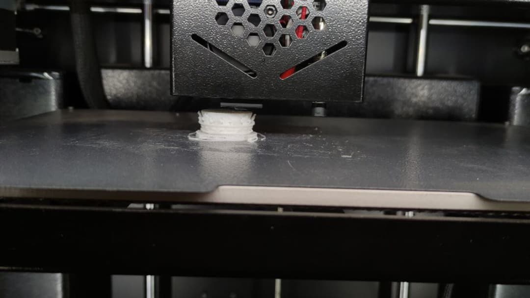A 3D printer's black print head lowers over a dark bed, building up a small white object layer by layer.