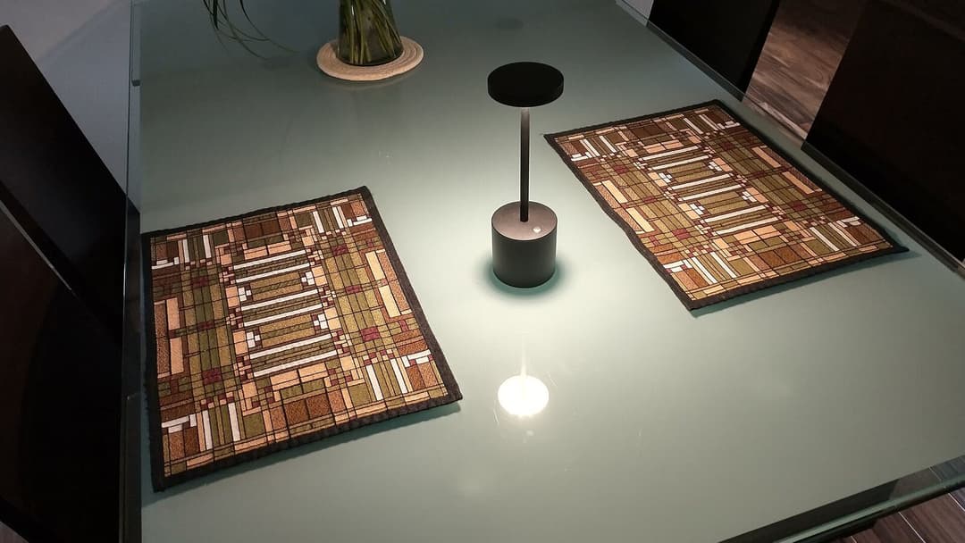 A small black cordless table lamp stands on a frosted glass tabletop between two patterned placemats, its light bouncing back off the glass as a bright spot of glare.
