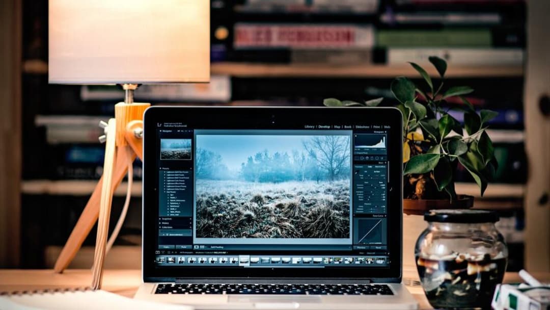 An open laptop on a wooden desk shows a photo-editing program, lit from the left by a wooden tripod desk lamp with a fabric shade, a leafy plant and a glass jar to the right, and rows of books on the shelves behind.