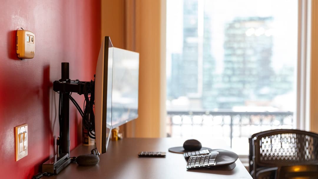 A monitor on an arm stands side-on at the end of a desk, at right angles to a tall window looking out on city towers, with a split keyboard, a mouse and an office chair in front of it.