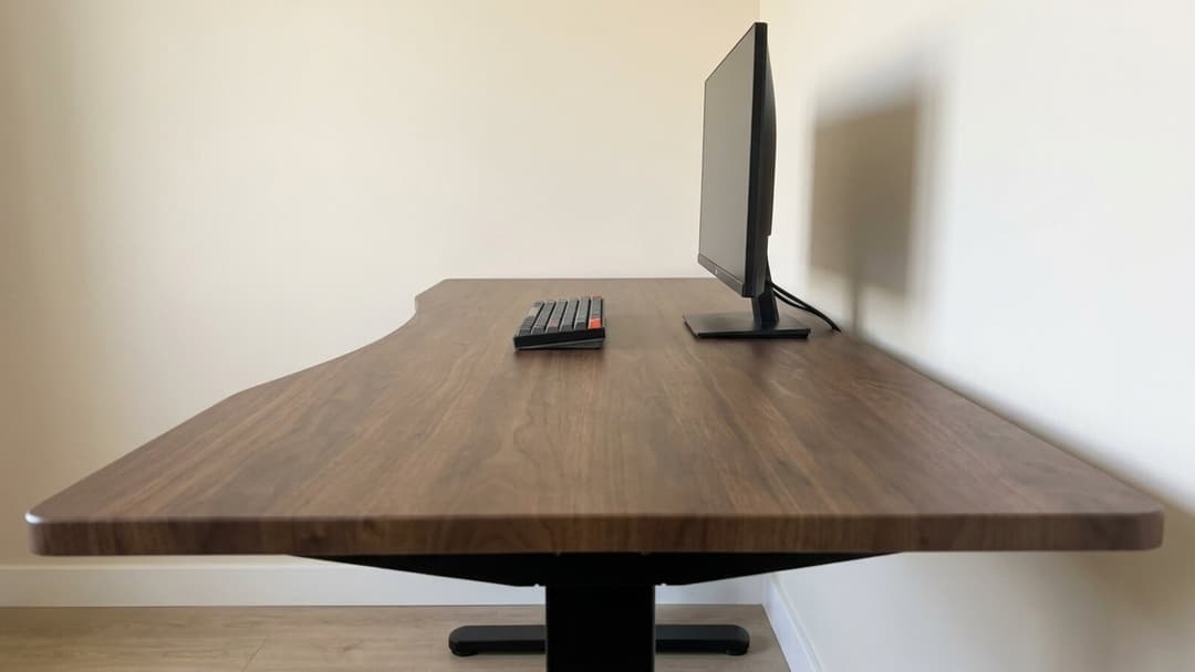 A walnut desk top sweeping away from the viewer to a monitor standing at the far back edge, a black keyboard set midway across, with a broad stretch of empty matte surface between the front edge and the screen.
