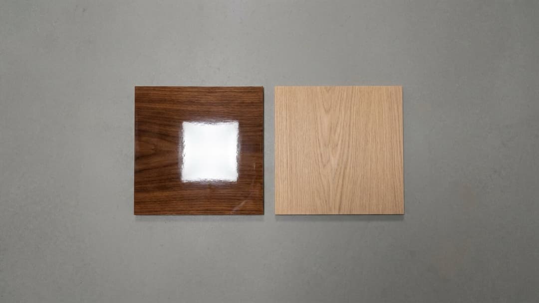 Two square wood swatches on a grey background: the darker glossy swatch on the left throws back a hard, bright square reflection of the light, while the paler matte swatch on the right scatters the same light evenly with no hotspot.