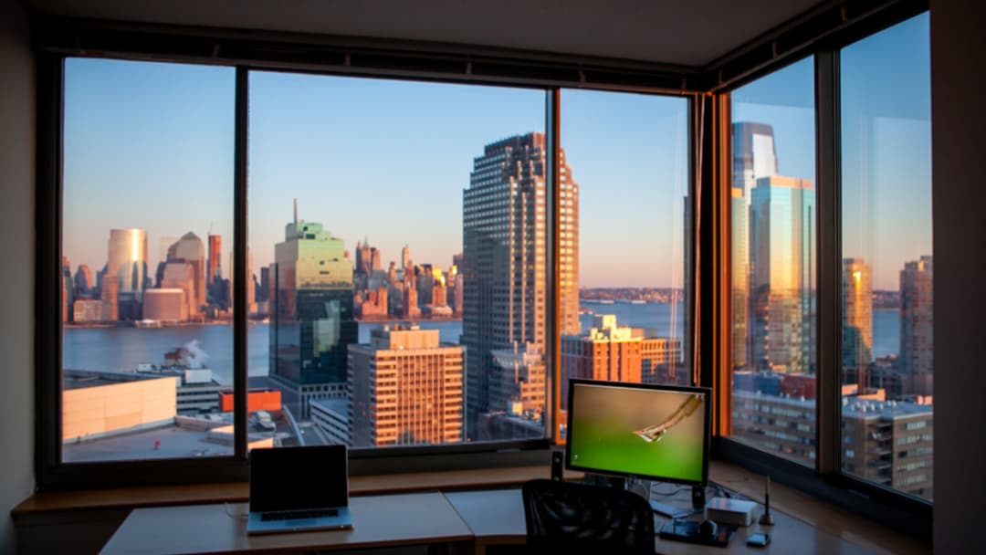 A home desk at dusk with a laptop and an external monitor and a dark office chair beside large corner windows over a city skyline.