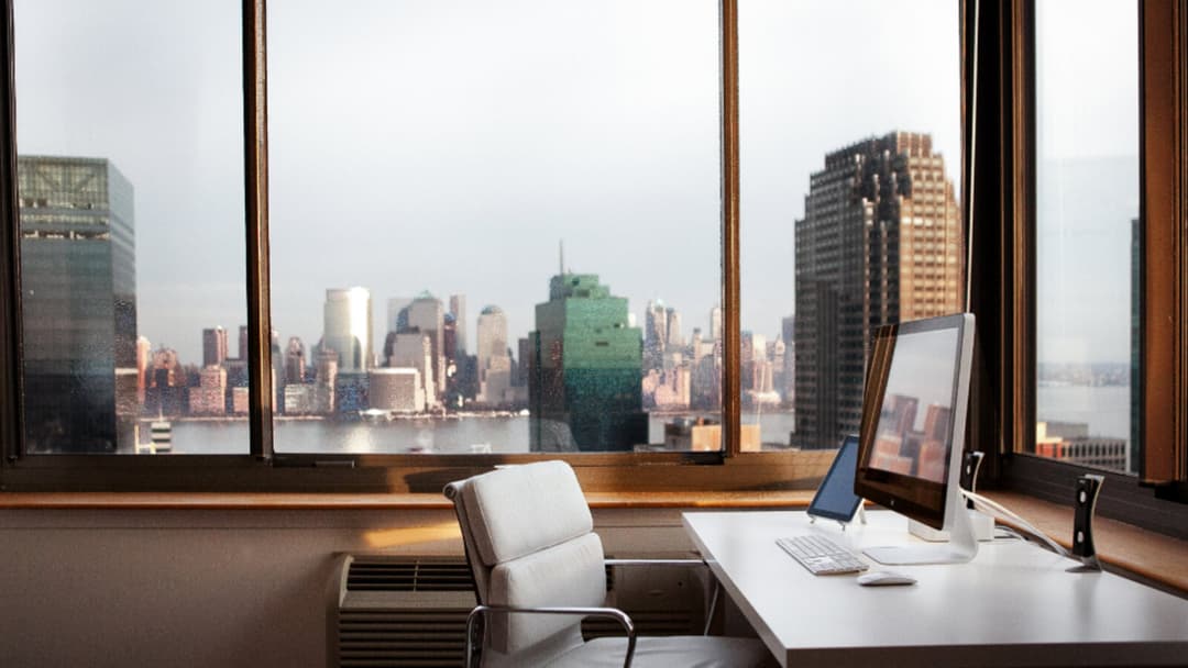 A white cushioned office chair with a chrome frame pulled up to a white desk by tall windows overlooking a hazy city skyline.