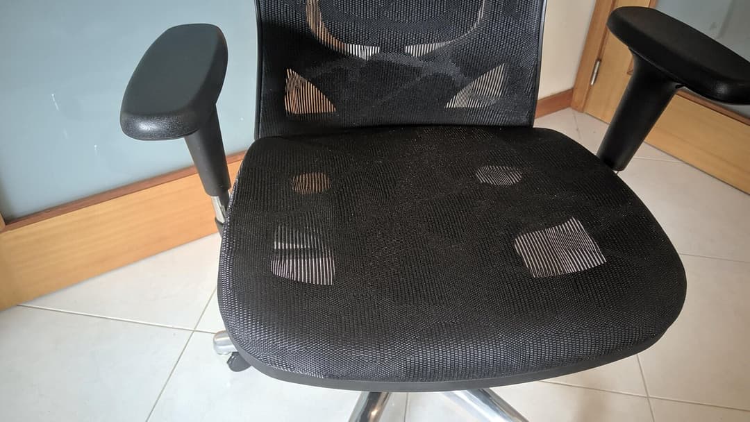 Close view of the black mesh seat and two padded armrests of an office chair on a tiled floor.