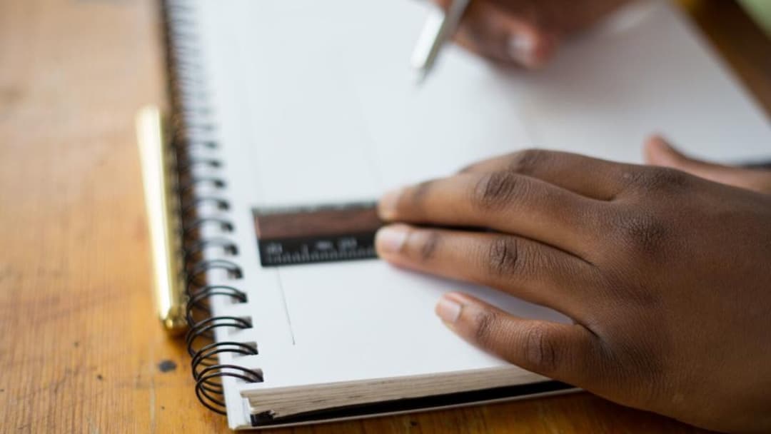 A hand resting on a spiral-bound notebook with a small black ruler across the page and another hand holding a pen to write, on a warm wooden desk.