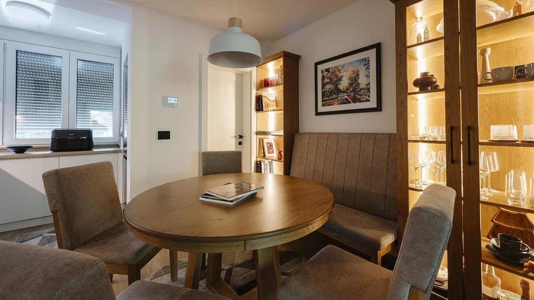 A round wooden dining table ringed by grey cushioned chairs and a padded bench, beside a lit glass display cabinet of glassware, under a white pendant lamp in a bright room.