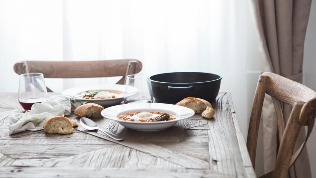 A rustic whitewashed wooden dining table set for a meal, with two white bowls of tomato stew, torn bread, a glass of red wine and a black pot between two wooden chairs by a bright curtained window.