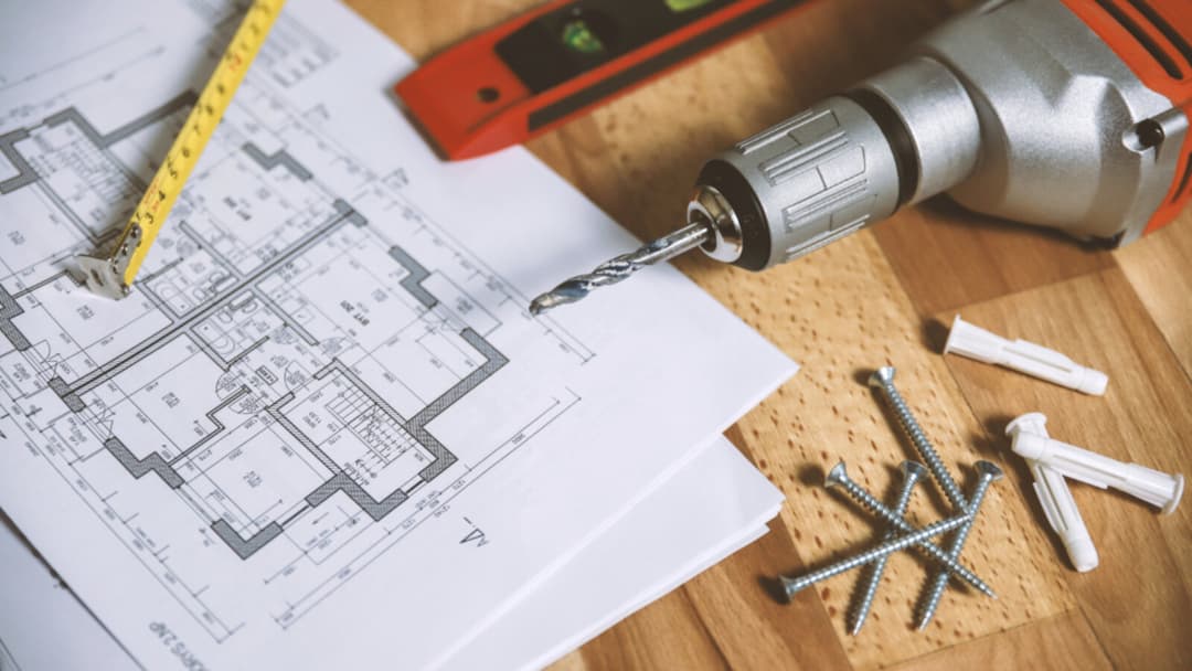 A yellow tape measure laid across a printed floor plan beside a red spirit level, a power drill, loose screws and wall plugs on a wooden surface.