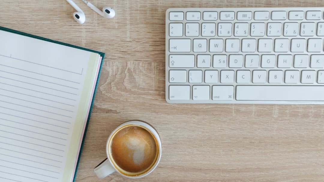 An overhead view of a light wooden desk with an open blank lined notebook, a cup of coffee, a pair of white earphones and a white keyboard.