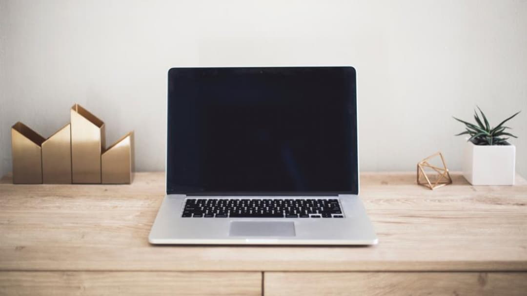 An open silver laptop with a blank screen on a light wooden desk, a gold magazine holder to the left and a small potted plant and a gold geometric ornament to the right, against a plain white wall.
