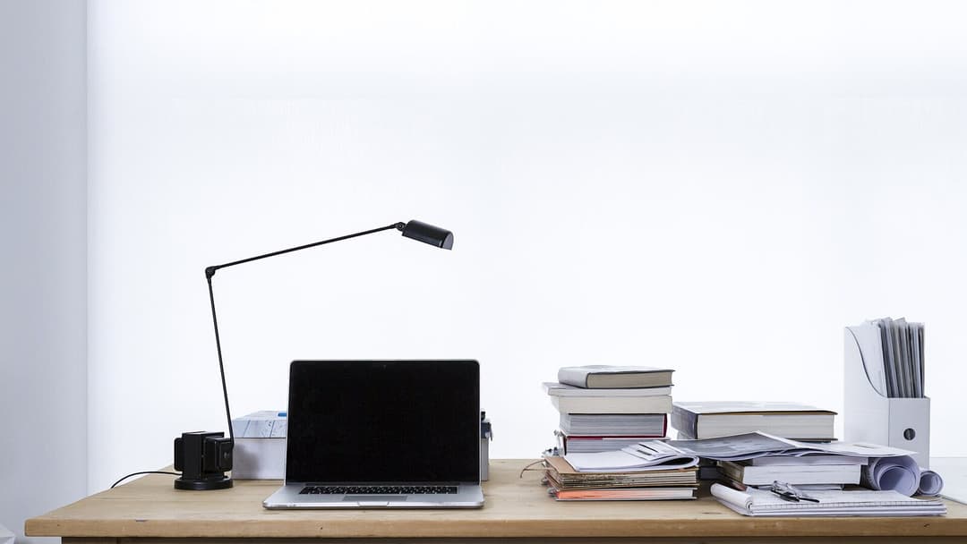 A light wooden desk against a white wall holding a black laptop with its screen off, a black anglepoise desk lamp, and a large pile of hardcover books, loose papers, notebooks and a white magazine holder stuffed with documents.