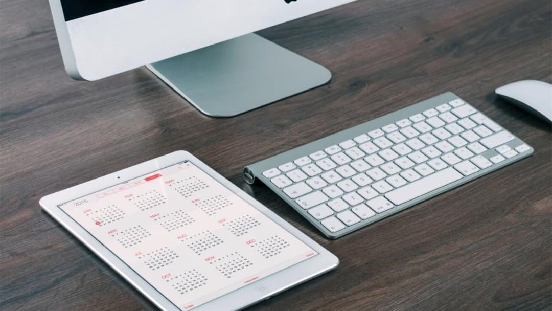 A slim silver external keyboard, a white mouse and a tablet showing a calendar on a dark wooden desk, below a monitor.