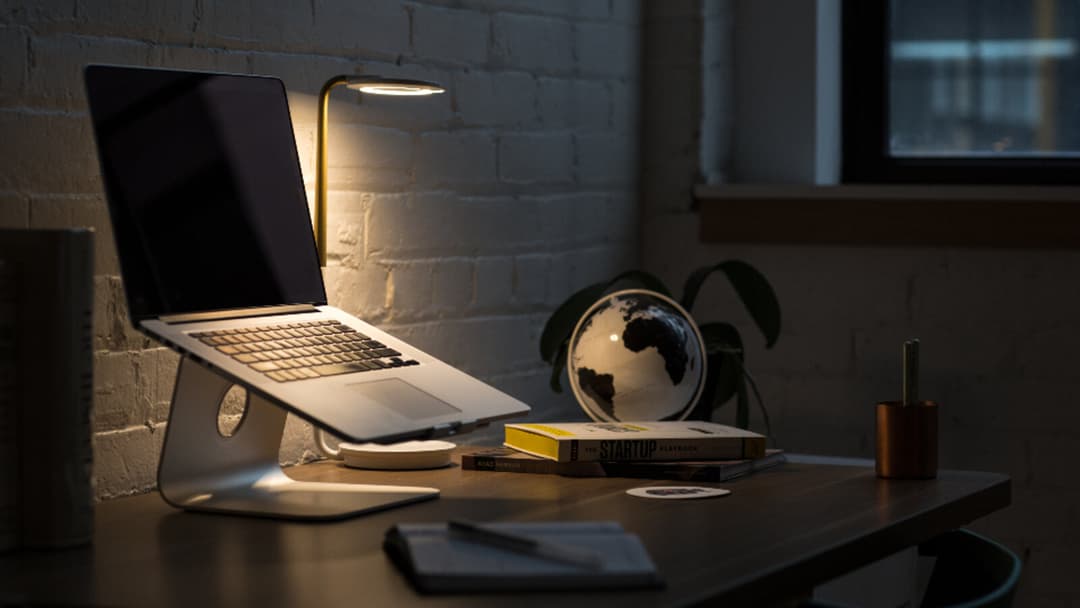 A laptop propped open on a curved aluminium stand on a dark wooden desk, lit by a desk lamp, with a globe and a stack of books beside it.