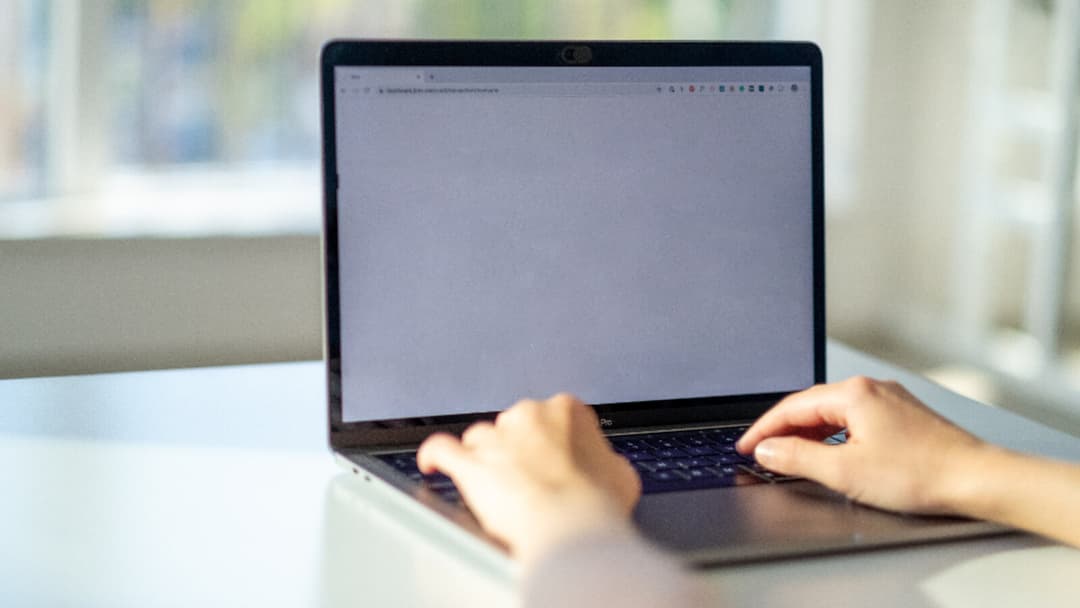 Two hands typing on a laptop that rests flat on a white desk, the screen low in front of a bright window.