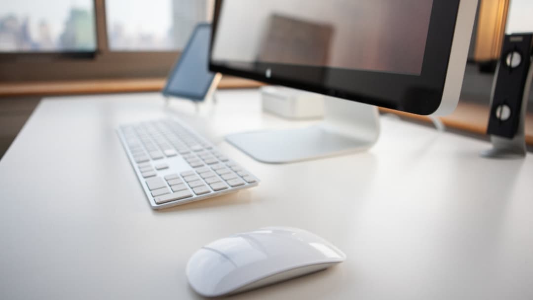 An all-in-one computer, a slim keyboard and a white mouse on a bright white desk beside a window.