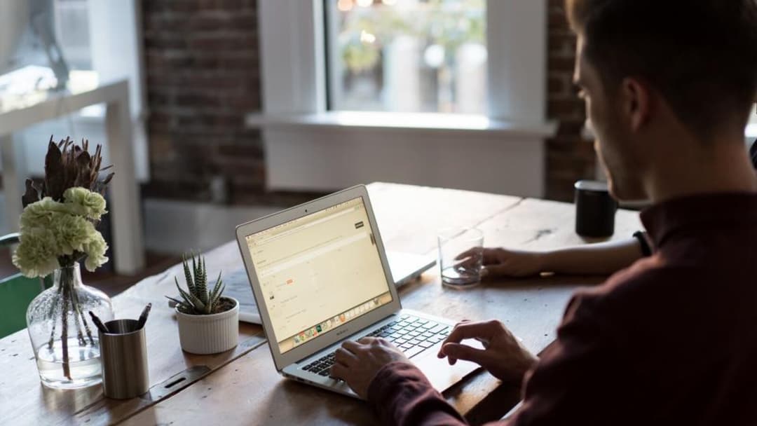 A person works at a laptop resting flat on a wooden table, the screen low and the head tipped down.