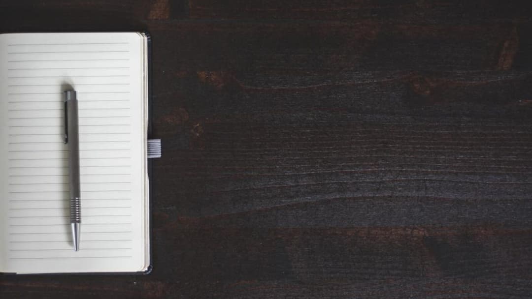 An open lined notebook with a slim metal pen resting on the page, on a dark wooden desk with a wide stretch of empty surface beside it.