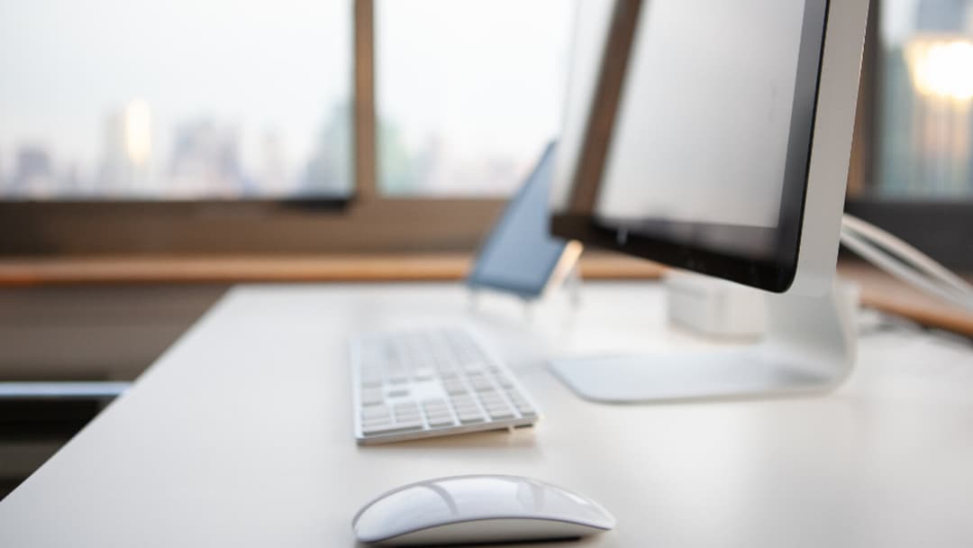 A silver all-in-one computer on a white desk beside a window with a blurred city skyline, a slim wireless keyboard laid in front of it and a white wireless mouse in the foreground.