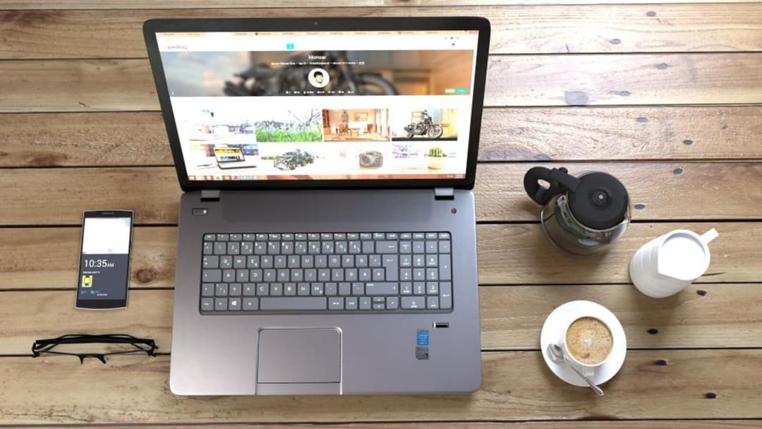 An overhead view of a laptop on a wooden plank table, with a smartphone, folded eyeglasses, a stovetop coffee pot, a small jug and a cup of coffee arranged around it.