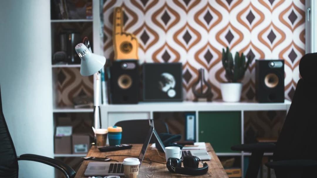 A wooden desk holding an open laptop, over-ear headphones, a coffee cup and a desk lamp, with a black office chair on each side and a white shelf of speakers, a cactus and ornaments against brown-and-white patterned wallpaper.