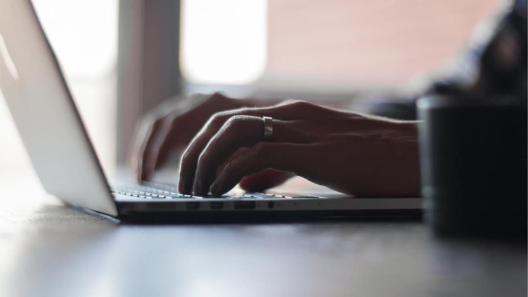 A side view of two hands typing on a laptop keyboard on a desk, backlit by a window, with no face visible.
