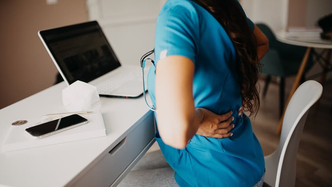 A person seated at a white desk with a laptop, pressing a hand to their lower back, with their face not visible.