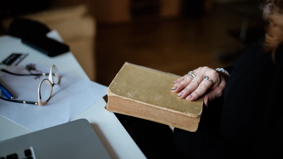 A hand resting on a worn old hardcover book beside reading glasses and a laptop on a desk, with no face visible.