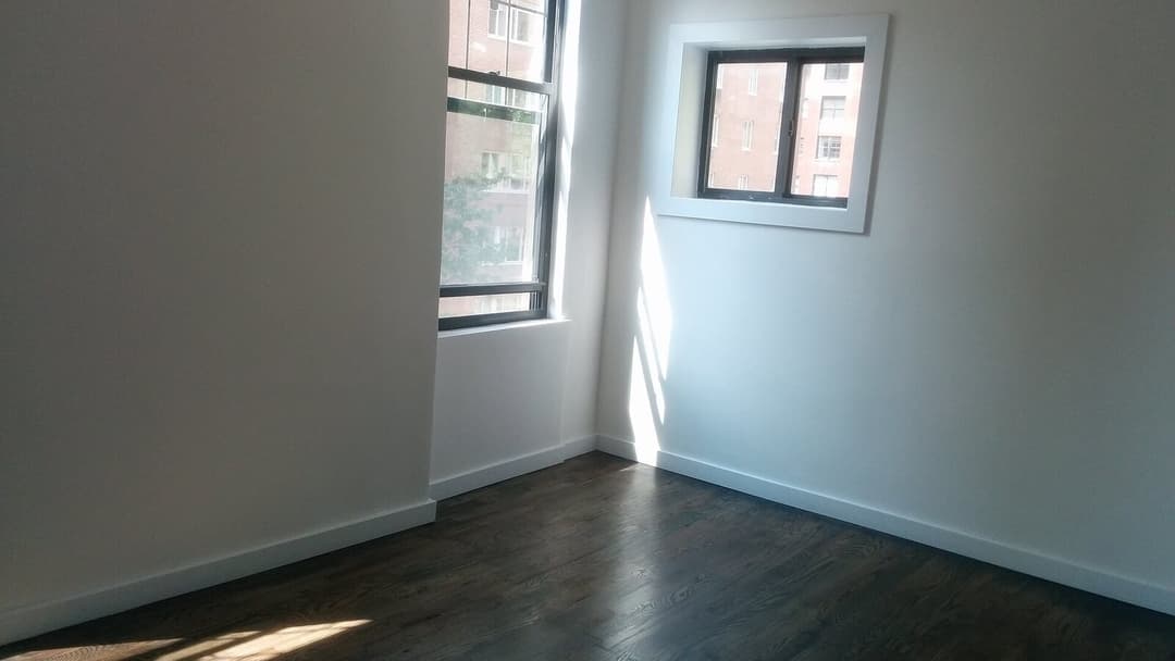 An empty room with a dark wooden floor and white walls, lit by a tall window and a small square window in the corner that look out onto a neighbouring building.