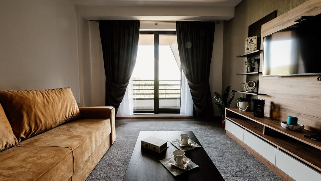 A modern living room with a tan suede sofa, a grey rug and a dark wooden coffee table holding a book and two white cups, in front of a glass balcony door framed by dark curtains, with a wall-mounted television above a wooden media console.