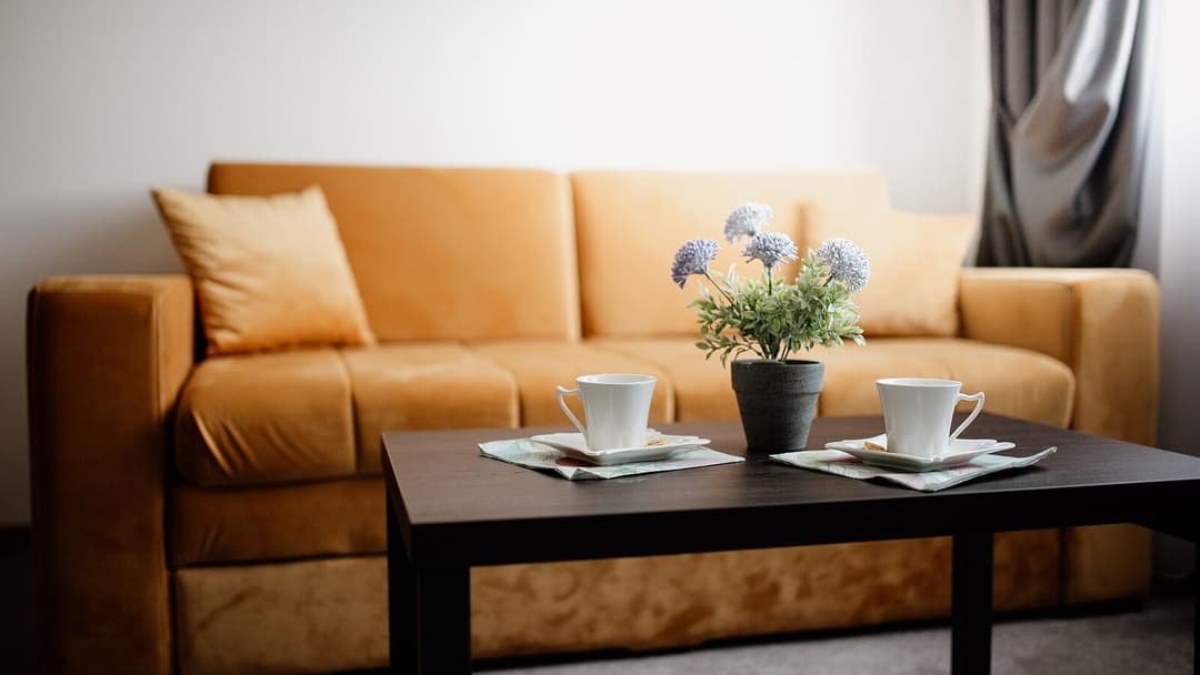 A tan velvet three-seat sofa with a matching cushion, behind a dark wooden coffee table set with two white cups on saucers and a small potted plant.