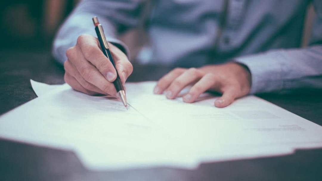 A person in a light blue shirt writing with a pen on a sheet of paper at a dark table, their other hand resting on the page.