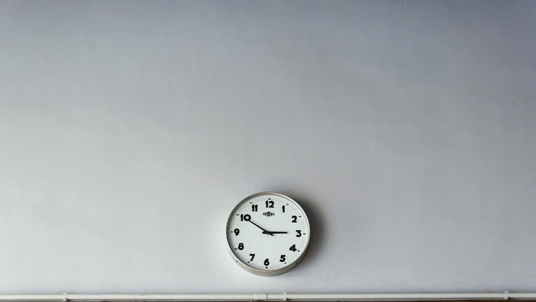 A plain round wall clock with a metal rim and a white face showing the numbers one to twelve, mounted on a pale grey wall.