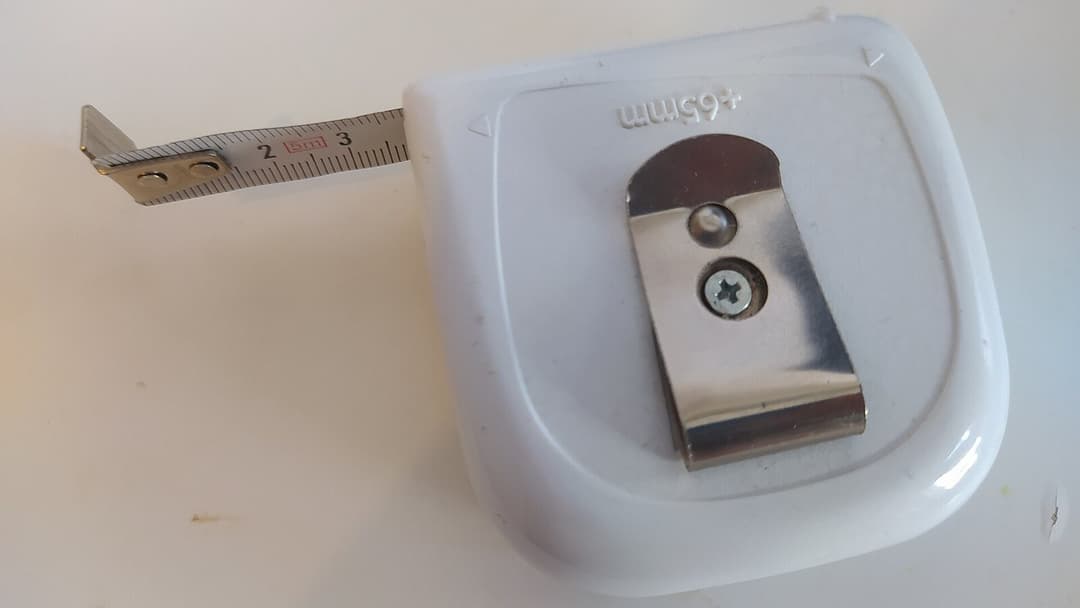 A white pocket tape measure on a pale table, its steel blade pulled out past the 2 and 3 centimetre marks.