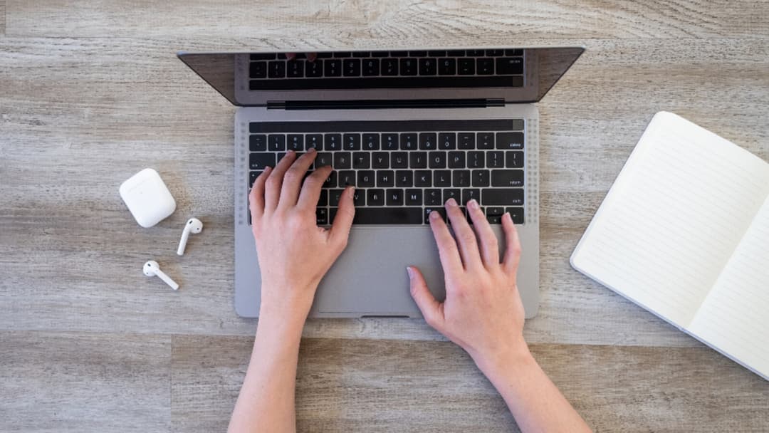 Overhead view of two hands typing on a grey laptop on a pale wood surface, with wireless earbuds in an open case to the left and a blank open notebook to the right.