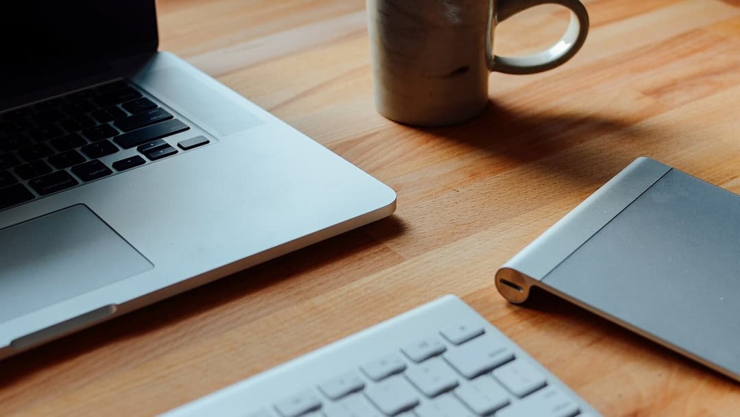A laptop, a ceramic mug, a slim wireless keyboard and a trackpad arranged on a warm honey-coloured wooden desk.