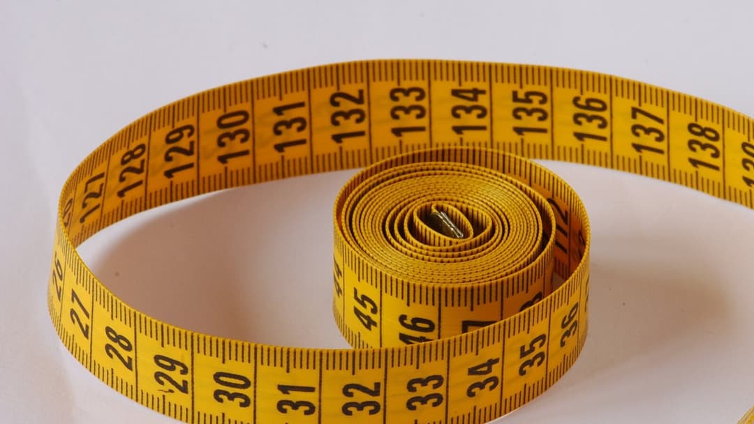A yellow tape measure loosely coiled on a pale surface, with its centimetre numbers from the twenties and the one-hundred-thirties printed along it.