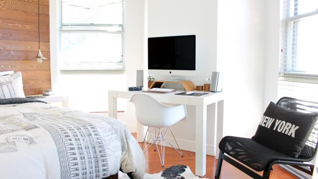 A white moulded chair pulled up to a white desk in a bright bedroom, with a monitor on a wooden stand and speakers on the desktop and a bed beside it.