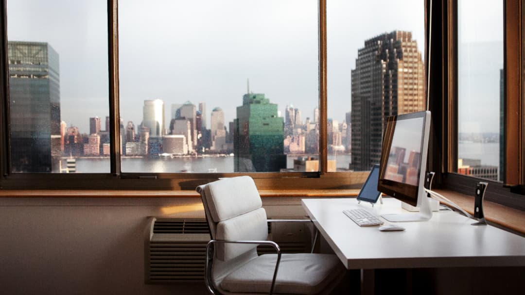 A white leather office chair with chrome arms beside a white desk under a wide window, a monitor and keyboard on the desktop and a city skyline beyond.