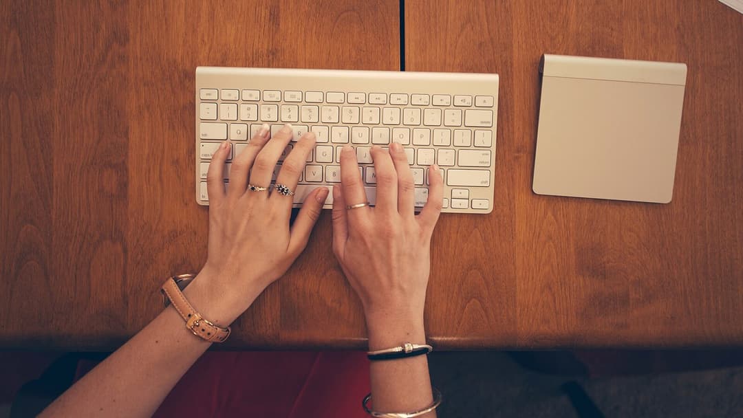 Two hands typing on a slim white keyboard laid on a warm wooden desk, seen from above, with a trackpad beside it.