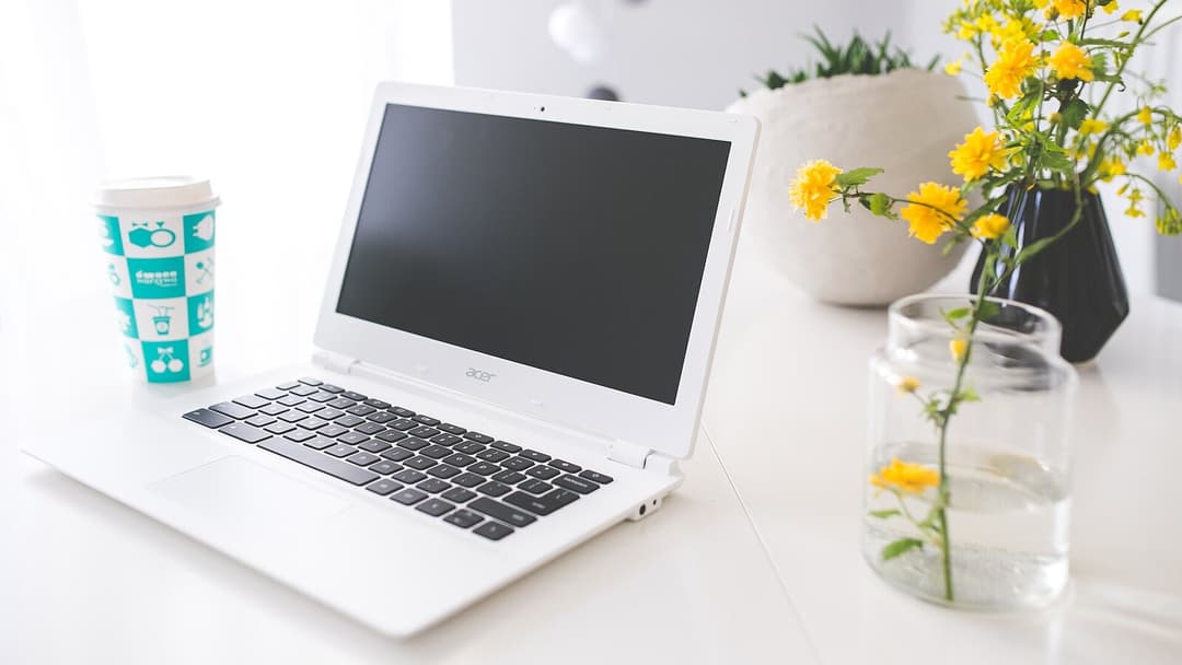 A white laptop with a blank dark screen on a bright white desk, a takeaway coffee cup with a teal pattern standing to its left and a clear glass jar and a dark vase of yellow flowers to its right.