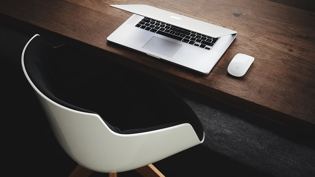 An empty white moulded chair with wooden legs pushed up to a dark wooden desk seen from above, an open silver laptop and a small white mouse resting on the desktop, with a grey floor below.