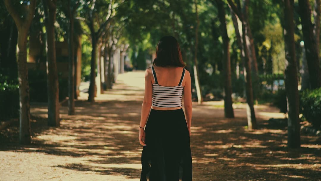 A woman seen from behind walking away down a sunlit, tree-lined gravel path, wearing a striped top and a long dark skirt, with rows of trees casting dappled shadows across the ground on both sides.