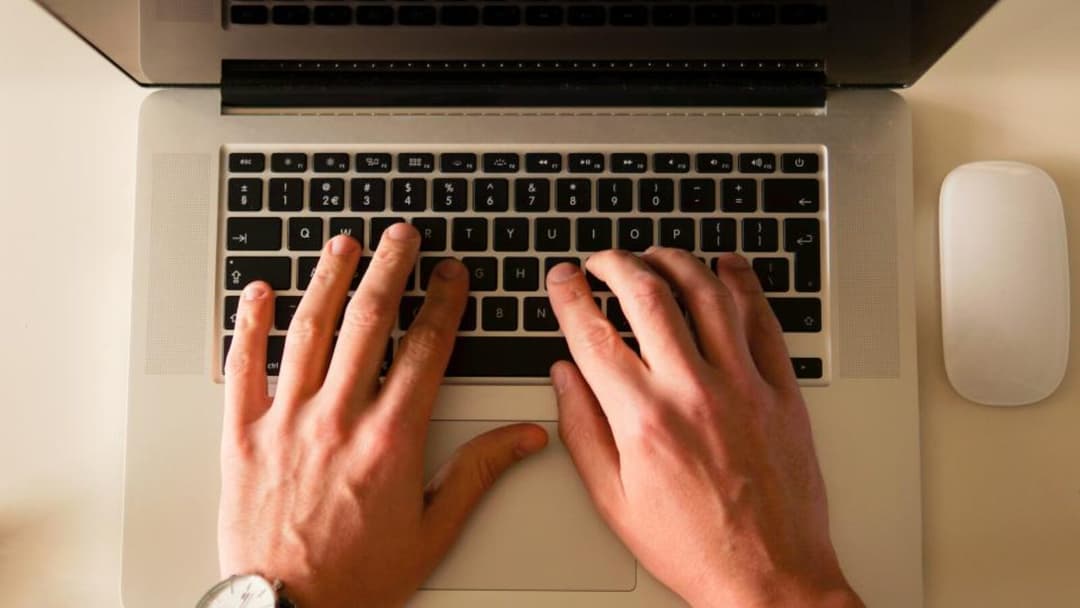 Two hands seen from directly above typing on a silver laptop's black keyboard, a white wireless mouse resting on the pale desk to the right and a wristwatch on the left wrist.