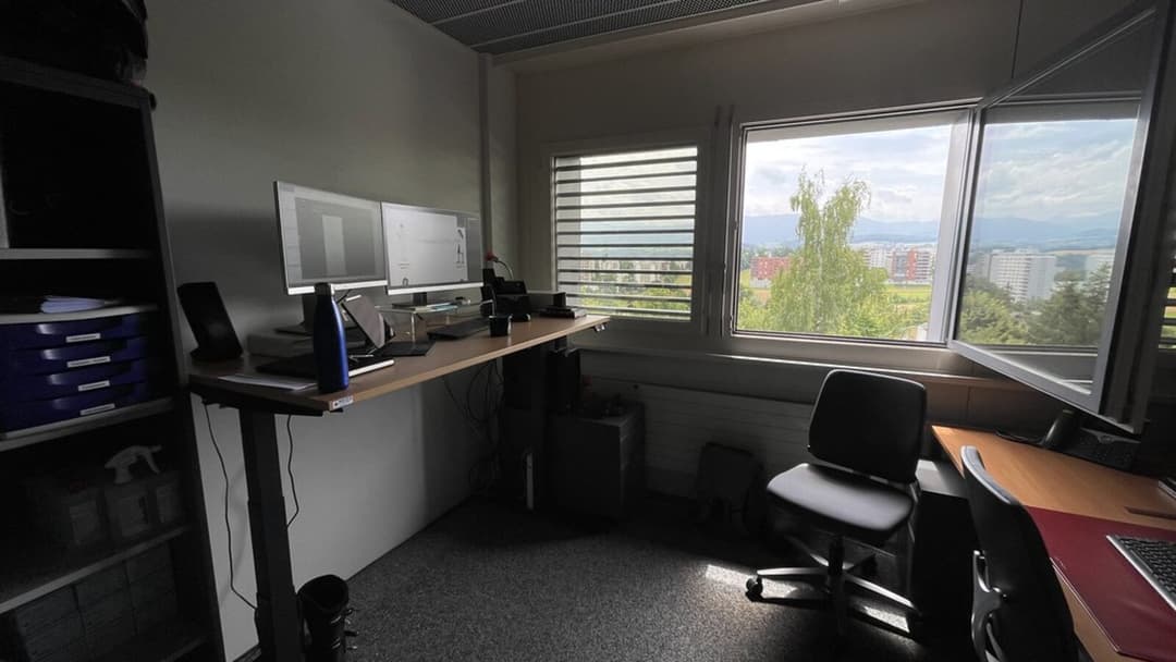A corner office with a long wooden-topped height-adjustable desk raised to standing height along the left wall, carrying two monitors, a shelf of blue box files beside it, a dark office chair and a second desk by large open windows that look out over trees, low buildings and distant hills.
