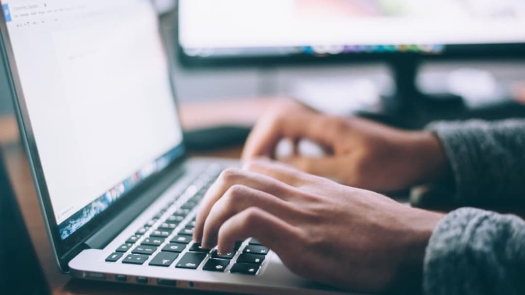 A side view of two hands typing on a laptop at a desk, the person's grey-sleeved arm reaching in from the right and a second monitor sitting blurred in the background.