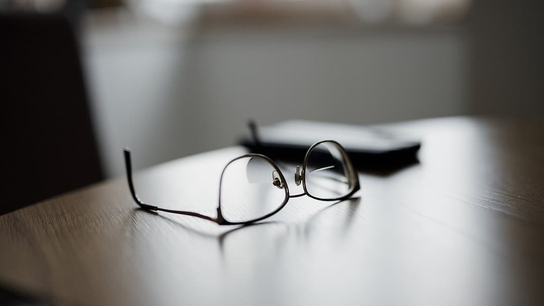 A pair of thin-framed reading glasses lying folded on a wooden table in soft daylight, with a dark notebook blurred behind them.
