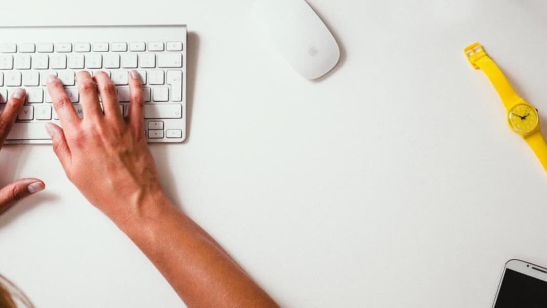 A top-down view of a hand resting on a slim white keyboard with the forearm flat on a white desk, a mouse, a yellow wristwatch and a phone edge nearby.