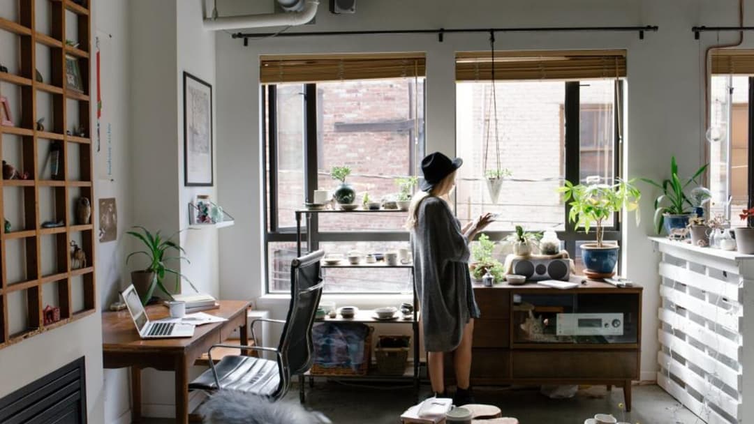 A sunlit loft home workspace with a wooden desk, an open laptop and a black leather office chair by tall windows, potted plants on a sideboard, and a person standing with their back to the camera.
